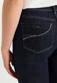 Jeans de mezclilla azul oscuro mostrados de lado, con un bolsillo trasero con costura curva y una textura suave.