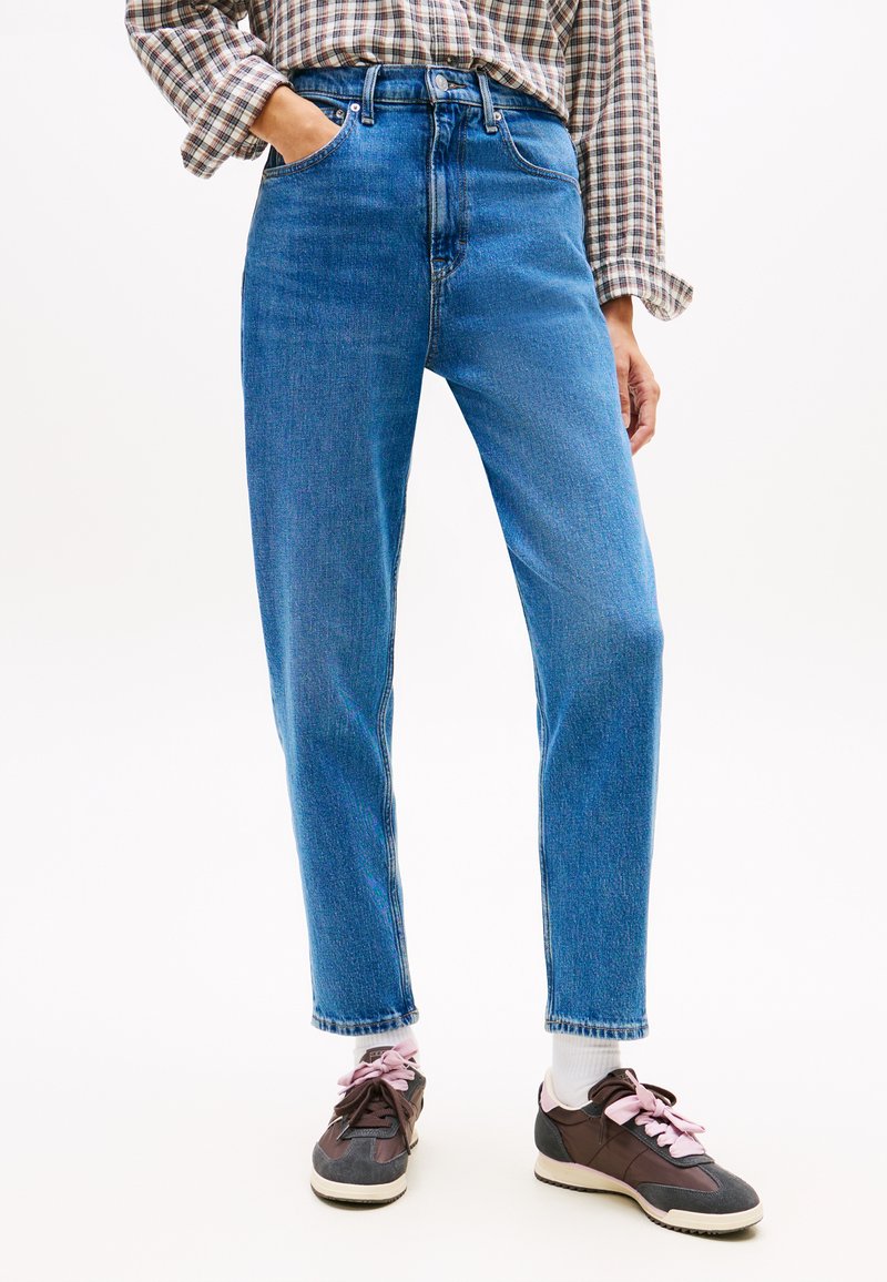 Persona che indossa jeans blu a vita alta e gamba dritta, camicia a quadri a maniche lunghe infilata, calzini bianchi e sneakers marroni con lacci rosa.
