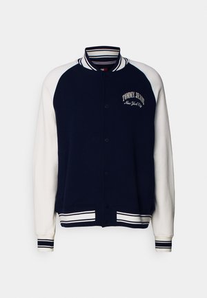 Laivastonsininen ja valkoinen yliopistotakki, jossa on raidallinen kaulus, hihansuut ja helma. Rinnassa teksti "Tommy Jeans New York City".