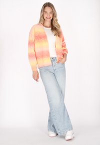 Överdimensionerad cardigan i gradientfärger av orange och rosa, bärs över en vit topp, parat med högmidjade ljusblå jeans och vita sneakers.