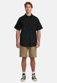 Camisa negra de manga corta con patrones geométricos dorados, botones en la parte delantera y un bolsillo en el pecho, combinada con pantalones cortos caqui y zapatillas negras.