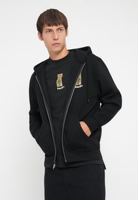Black zip-up hoodie with front pockets, textured fabric, worn over a black t-shirt featuring graphic prints in gold and white.