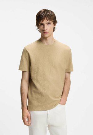 Jeune homme portant une chemise marron clair texturée à manches courtes et un pantalon blanc, debout avec une main dans la poche sur fond blanc.