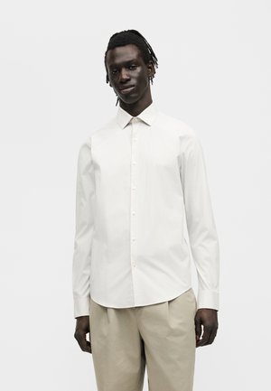 Άνδρας στέκεται με το πρόσωπο προς τα εμπρός, φοράει άσπρο πουκάμισο με μακριά μανίκια και μπεζ παντελόνι μπροστά σε απλό λευκό φόντο.