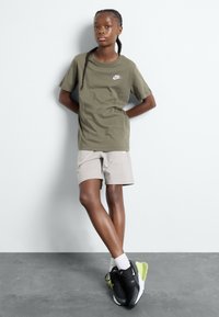 Olivno zeleno bombažno t-shirt s belim Nike logotipom, v kombinaciji s svetlo bež kratkimi hlačami in črnimi športnimi copati z limonastimi poudarki.