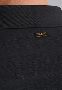Zwarte textuurstof met een geribbeld kraag, met een kleine gouden logo patch die "American Classic" en een vleugeldetail afbeeldt.