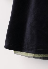 Black fabric, soft texture, featuring a layered design with a sheer yellow-green check pattern at the hem. Stitched edge detail visible.