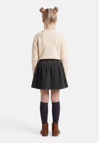 Enfant aux cheveux blonds coiffés en deux chignons, portant un pull crème, une jupe en velours côtelé noire, des chaussettes noires hautes jusqu'aux genoux et des bottes marron, debout de dos.