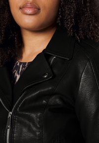 Svart imitation läderjacka med dragkedja och tryckknappar i kragen. Har strukturerade ytor och kontrasterande sömmar. Närbild.