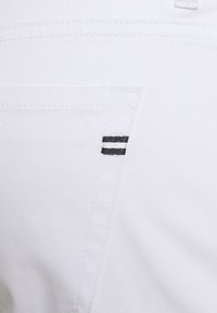 Close-up van witte stof die een gestikte zakrand toont met een klein zwart-wit dubbelstreepjes stoflabel.