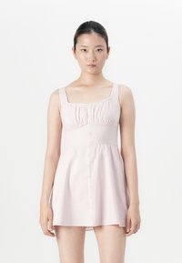 Giovane donna con i capelli scuri raccolti, che indossa un vestito smanicato di un rosa pallido con bottoni sul davanti, in piedi contro uno sfondo bianco.