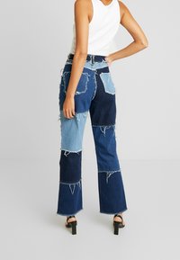 Kvinna som bär högmidjade patchwork denimjeans med fransade kanter, i kombination med en vit ärmlös topp och svarta klacksandaler.