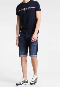 Námořnicky modré tričko s krátkým rukávem s bílými a červenými pruhy, spárované s tmavými denimo šortkami s ohrnutým lemem a bílými teniskami s olivovými akcenty.