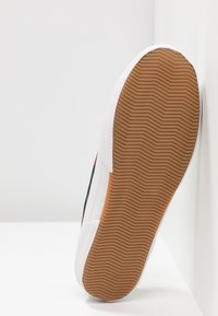 Pohja valkoisista lenkkitossuista, joissa on ruskea zigzag-kuvioinen kumipohja, kulmassa valkoista pintaa vasten.