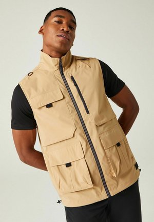 Uomo che indossa un gilet beige con più tasche sopra una camicia nera a maniche corte, in piedi davanti a uno sfondo chiaro uniforme.
