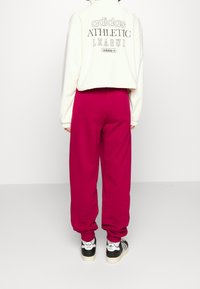 Person klädd i en vit Adidas sweatshirt med texten "ATHLETIC LEAGUE", ljusrosa sweatpants och svarta och vita Adidas sneakers som står.