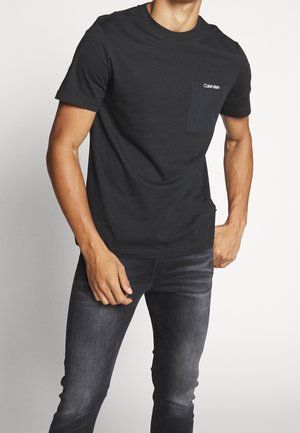 Homme portant un t-shirt noir uni Calvin Klein avec une poche poitrine et un jean délavé foncé, debout devant un fond blanc.