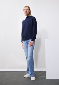 Sudadera azul marino con cuello alto, combinada con jeans acampanados de color azul claro y zapatillas blancas. Diseño sencillo, textura suave, fondo neutro.