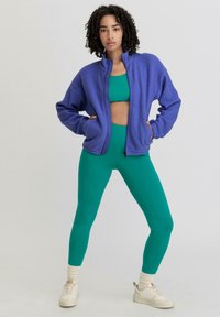 Paarse fleece zip-up jas met een hoge kraag, gecombineerd met strakke groene leggings. Model staat zelfverzekerd met de handen in de zakken.