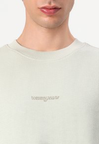 Lys grå, struktureret sweater med ribbet halsudskæring og "tommyjeans" logo broderet i en lidt mørkere nuance på brystet.