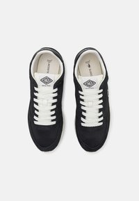 Black sneakers made of suede and mesh fabric, featuring white laces, a padded collar, and a logo patch on the tongue. Flat rubber sole.