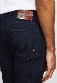 Homme portant un jean bleu foncé avec une poche arrière visible et un patch en cuir marron Tommy Hilfiger au-dessus de la poche.