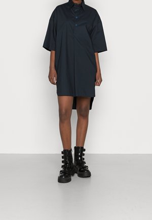 Person iført en løstsiddende sort kortærmet skjortekjole og sorte læderankelstøvler med flere spænderemme.