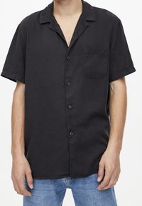 Chemise noire à manches courtes en tissu lisse, dotée d'un col cranté, d'une patte de boutonnage à l'avant et d'une poche poitrine unique.