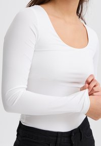 Fehér, hosszú ujjú felső mélyen kivágott nyakkivágással, rugalmas anyagból készült, testhez simuló ujjak és sima felület jellemzi. Fekete nadrággal viselve.