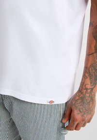 Bílý bavlněný tričko s krátkým rukávem, kombinované se zelenými a bílými pruhovanými kalhotami. Obsahuje malou značkovou etiketu na lemu.