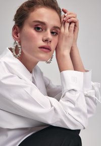 Blusa bianca con colletto, polsini abbottonati con ricamo e fascetta legata. Il modello indossa orecchini con grandi gemme trasparenti. Sfondo nero.