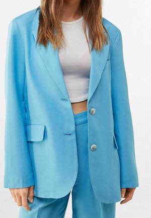 Donna con blazer azzurro chiaro e pantaloni coordinati, indossando un top corto bianco, mostrata dalle spalle fino a metà coscia su uno sfondo bianco.