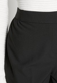 Černé látkové kalhoty s širokým pasem, s plisovaným designem a hladkou texturou, v kombinaci s bílým topem s žebrovanou strukturou.