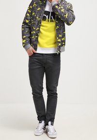 Chaqueta bomber gris con patrón de camuflaje amarillo sobre un sudadera amarilla y blanca, combinada con jeans negros y zapatillas blancas.