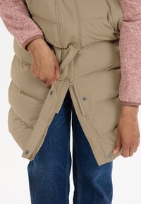 Beige pufferveste med lynlås og trykknapper, båret over en lyserød tekstureret langærmet top. Hænderne justerer lynlåsen.
