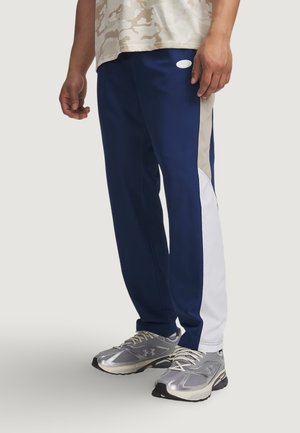 Uomo che indossa pantaloni della tuta blu navy con pannelli laterali beige e bianchi e scarpe da ginnastica argento, in piedi davanti a uno sfondo semplice.