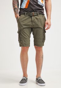 Bermudas cargo verde olivo con múltiples bolsillos laterales, un corte relajado y un cinturón de tela. Combinadas con zapatos casuales grises y una camisa oscura.