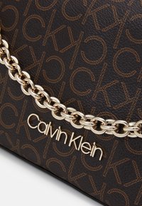 Ruskea käsilaukku, jossa on painettu CK-kuvio, kullanvärisellä ketjuolkaimella ja kultaisella Calvin Klein -logolla. Karkeapintainen nahkamateriaali.