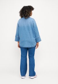 Casaco de ganga azul com um corte descontraído, mangas longas e uma bainha arredondada. Combinado com jeans azuis flare e ténis brancos.