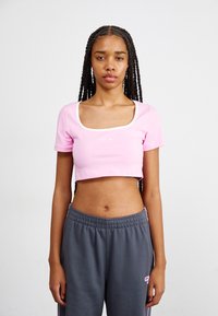Krátký růžový crop top se čtvercovým výstřihem a bílým lemem. Uprostřed se nachází malé bílé logo Nike. Kombinováno se šedými tepláky.