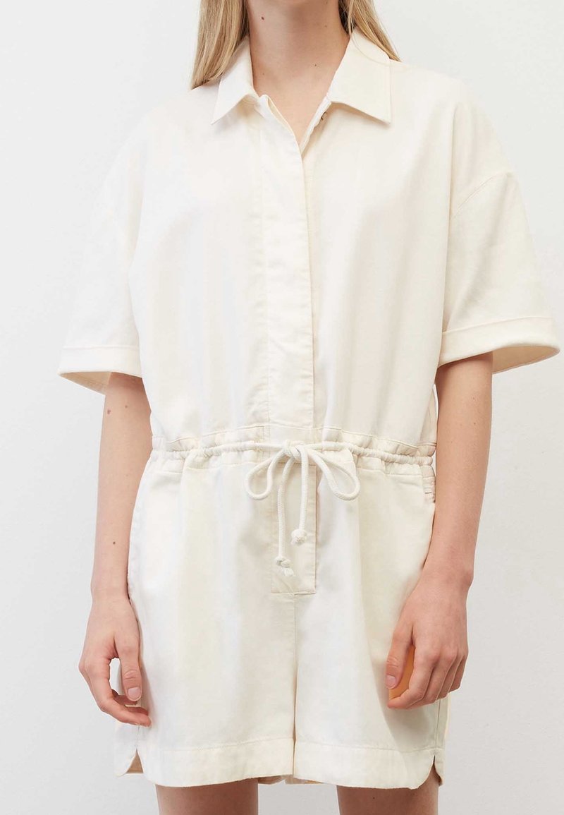 Femme portant une combinaison ample crème à manches courtes, avec col et taille ajustable par cordon, debout devant un fond uni.
