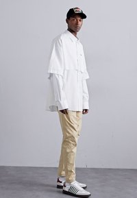 Weißes, übergroßes Hemd, beige Hose, grafische schwarze Mütze und klassische weiße Sneaker mit grünen und roten Akzenten. Glattes Material, lockere Passform.