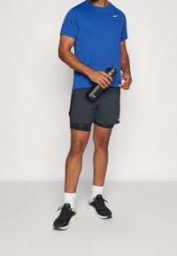 Camiseta deportiva azul con logo de Nike, combinada con pantalones cortos negros con forro de malla. Botella de agua negra en la mano, zapatillas de correr negras con suelas blancas.