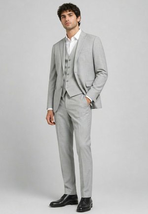 Homme portant un costume trois pièces gris clair avec des chaussures noires, debout avec une main dans la poche devant un fond gris uni.