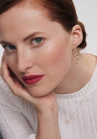 Złote serduszka w kształcie kolczyków o kaskadowym wzorze, noszone przez modelkę w lekkim swetrze i delikatnym czerwonym odcieniu ust.