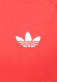 Raudonas medvilnės audinys su baltu siuvinėtu trefoil logotipu, turinčiu tris lapus ir du horizontalius dryžius po juo.