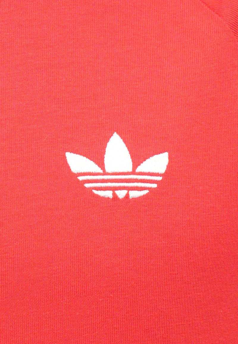 Un tessuto di cotone rosso presenta un logo a trifoglio ricamato in bianco con tre foglie e due strisce orizzontali sottostanti.