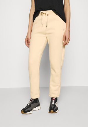 Persona con pantalones beige con cordón, camiseta negra y zapatillas blancas y negras, de pie contra un fondo claro y liso.