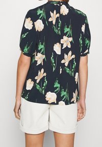 Květinová halenka s krátkým rukávem z tmavě námořnického materiálu, s bledě béžovými a zelenými květinovými vzory, s otvorem vzadu.