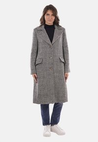 Cappotto in lana a spina di pesce grigio con due tasche frontali e bottoni. Maniche lunghe e colletto classico, abbinato a jeans blu e sneakers bianche.